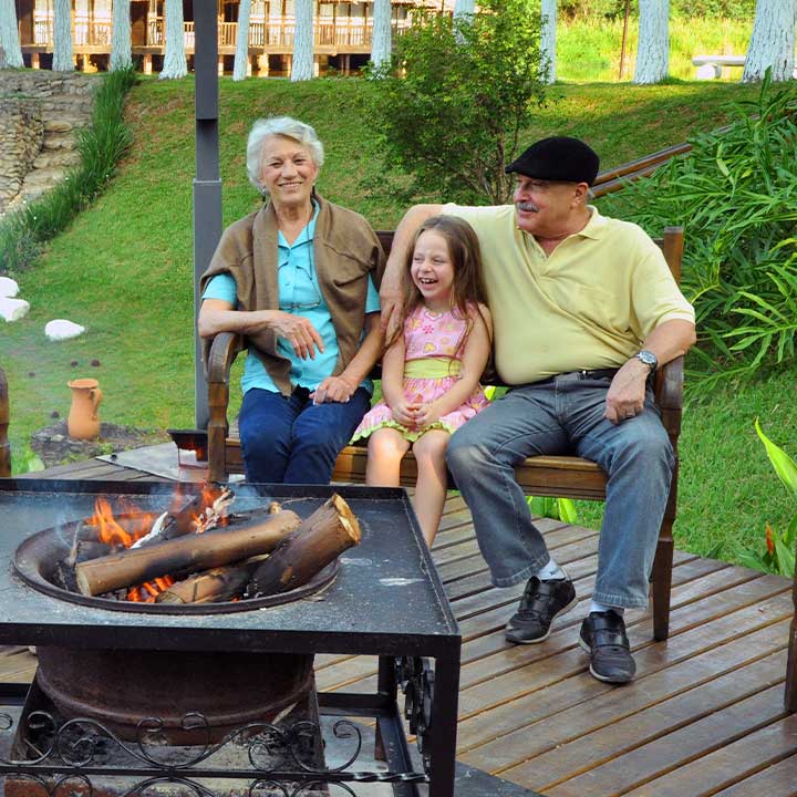 Imagem de uma família reunida ao redor de uma fogueira no jardim, sorrindo e aproveitando momentos de lazer ao ar livre com crianças e idosos.