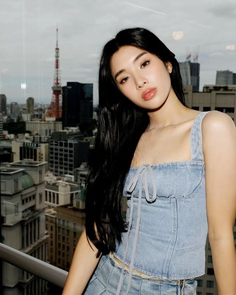 This contain KATSEYE Sophia   with long black hair standing in front of a cityscape and looking at the camera