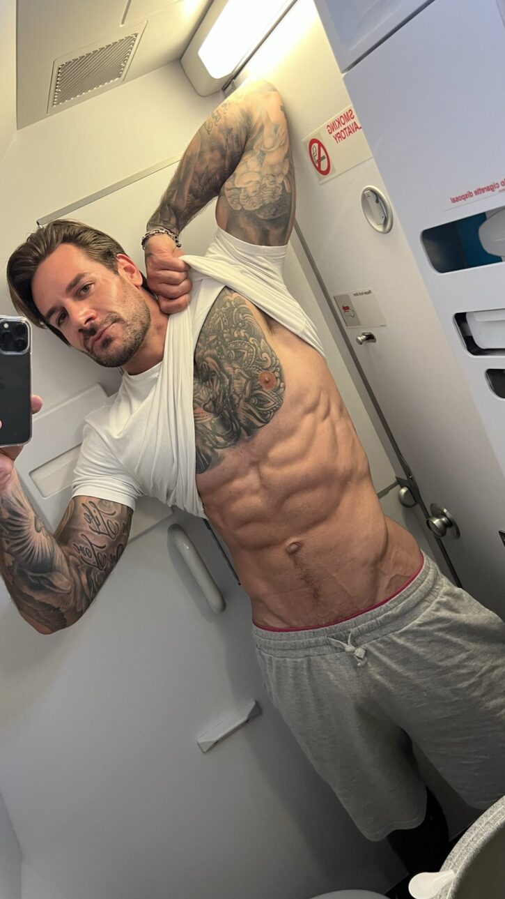 Papi Kocic taking an iphone mirror selfie in an airplane lavatory in grey sweatpants lifting up his white t-shirt to reveal his abs and tattoos