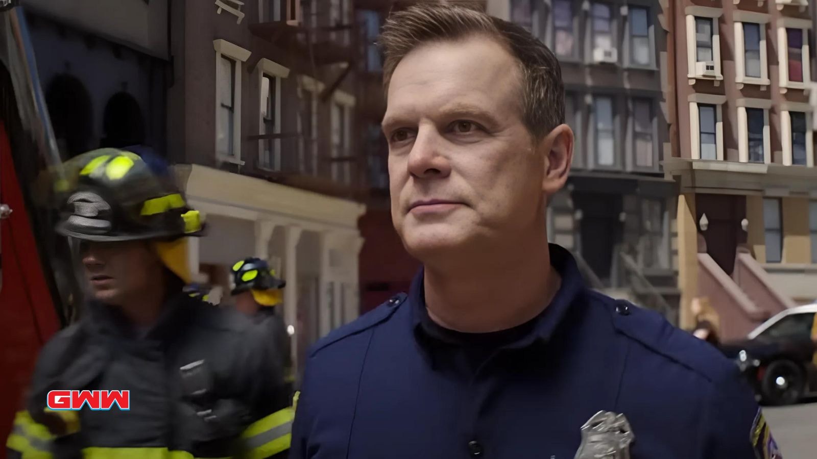 Bobby stands in front of a building with firemen behind him.