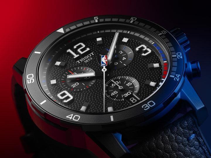 Close-up of the Tissot Supersport Chrono NBA Edition watch, featuring a textured black dial, bold numerals, chronograph sub-dials, and an NBA logo on the central second hand. The watch has a black bezel with white markings and red and blue accents, set against a dramatic red and black gradient background.