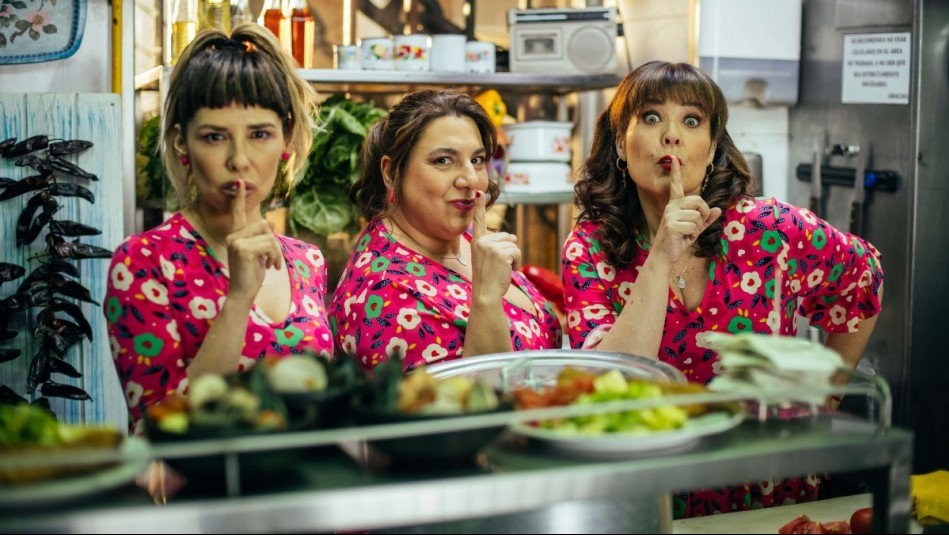 Tres mujeres en una cocina comercial, vestidas con atuendos coloridos con estampado de flores, haciendo el gesto de silencio con un dedo en los labios.