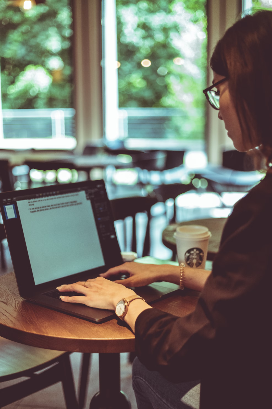  A woman wearing glasses sits at a table in a coffee shop and writes an academic paper on her laptop.