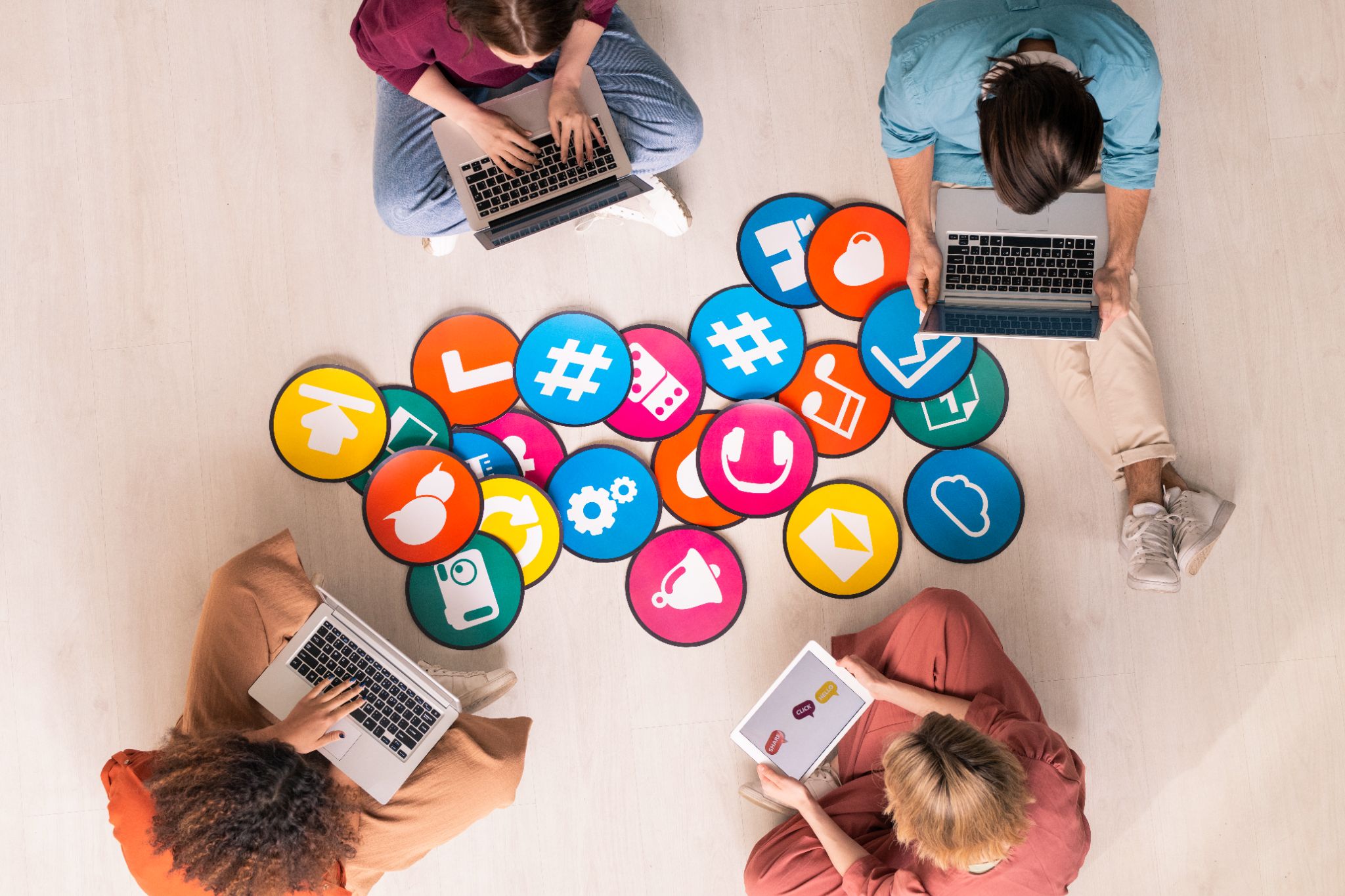 A group of young people sitting in a circle on the floor, using laptops and tablets, surrounded by colorful circular icons representing various digital and social media elements, including hashtags, music notes, and chat symbols