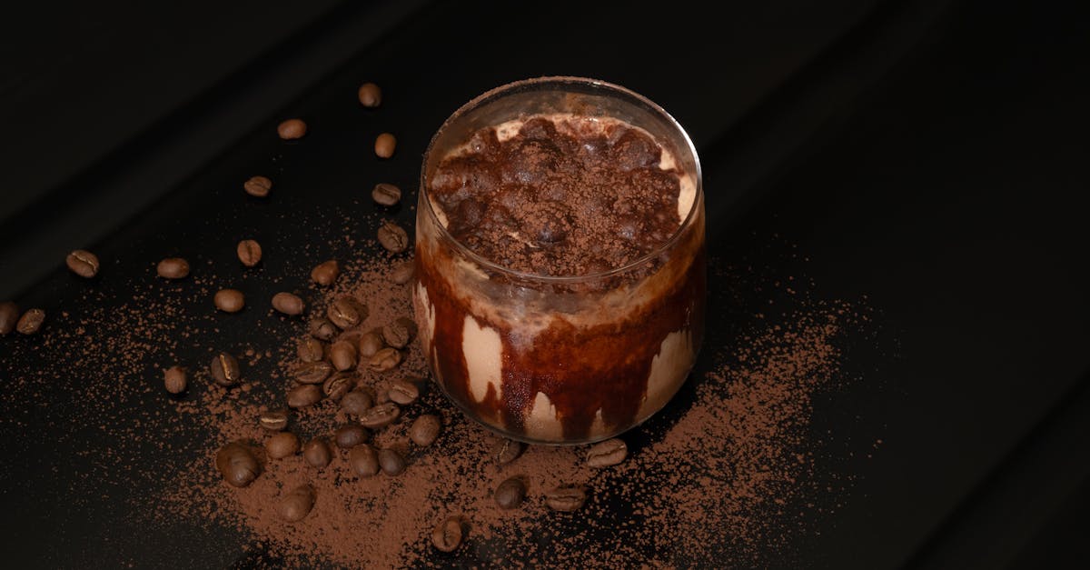 A tall glass filled with a dark brown, frothy beverage. The beverage is topped with chocolate syrup and cocoa powder, with a few coffee beans scattered around the base of the glass.