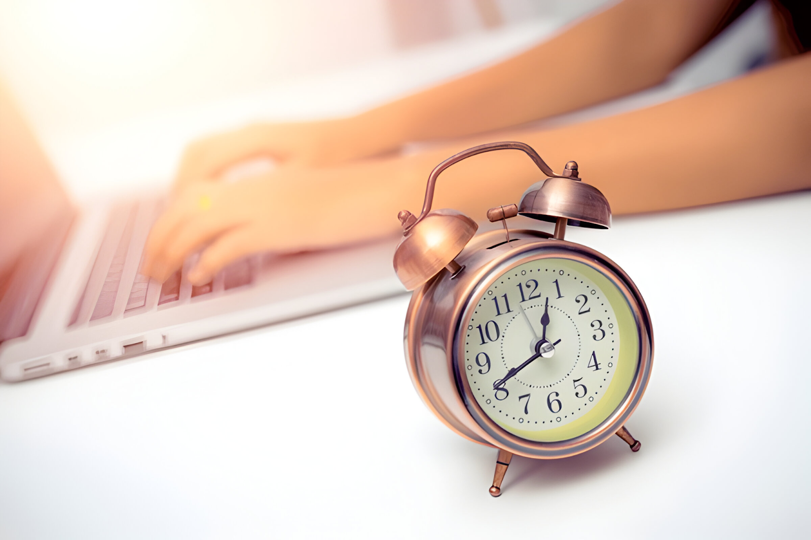 Close-up of an alarm clock on a desk with a blurred person typing on a laptop in the background, symbolizing time and effort savings in productivity.