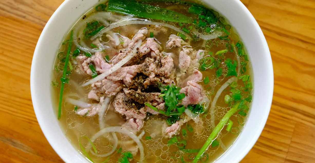 (Bowl of Vietnamese pho with rare sliced beef, onions, scallions, cilantro, and freshly cracked black pepper in a clear, aromatic broth.)