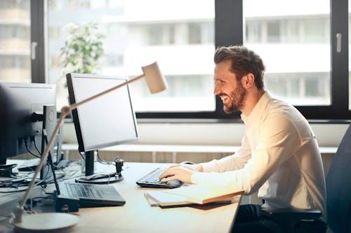 Free A man smiling while working at an office desk with a computer and natural daylight streaming in through large windows. Stock Photo