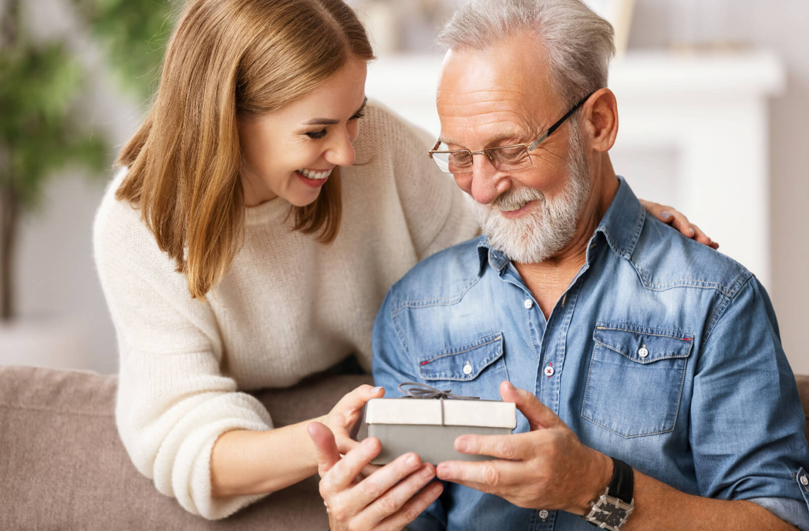 A young adult handing their smiling older parent with dementia a small gift while they both laugh.