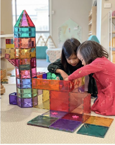 A pair of siblings are joyfully building a tall tower and structure using colorful magnetic tiles, demonstrating their creativity and teamwork. The older sibling teaches the younger one about critical problem-solving skills by placing larger tiles at the bottom for stability, ensuring their impressive architectural design stands strong as they work together with focus and enthusiasm.