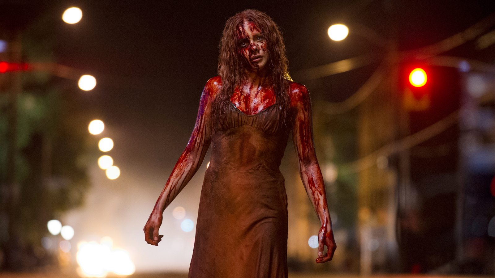 Mujer joven cubierta de sangre, vestida con un camisón oscuro y con una expresión inquietante, camina sola por una calle en la noche iluminada por luces de fondo borrosas. Su cabello desordenado y el escenario oscuro crean una atmósfera de terror y suspenso.