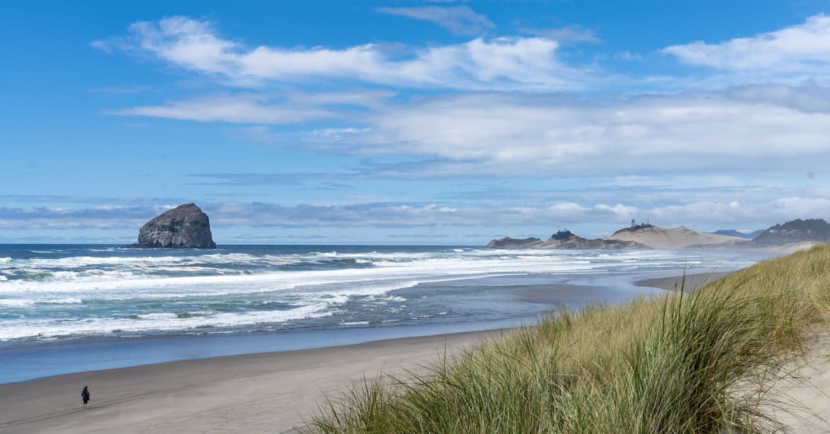 View of Haystack Rock at Cape Kiwanda from a sandy beach with grassy dunes on a cloudy day with blue skies