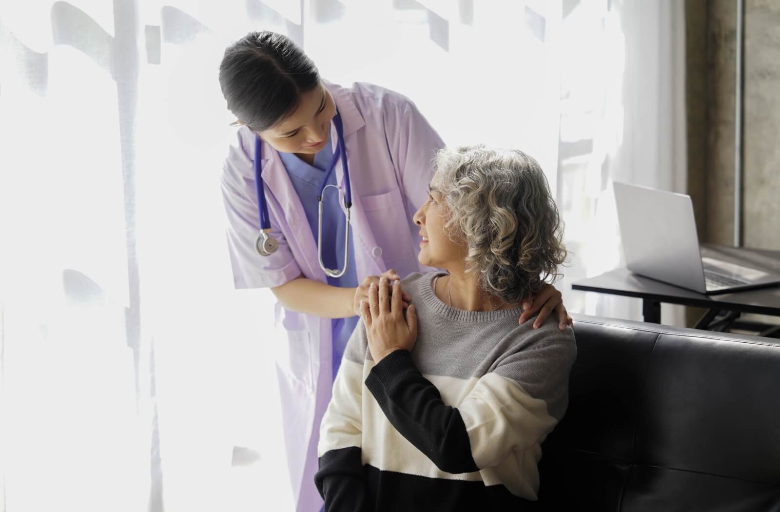 A caregiver with a stethoscope smiles while placing their hands on a senior resident's shoulder.