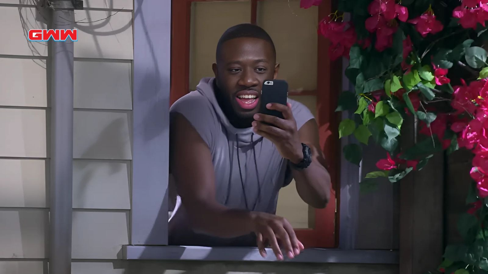 Malcolm (Sheaun McKinney) leaning out of a window with a phone, laughing amidst vibrant flowers in The Neighborhood Season 7