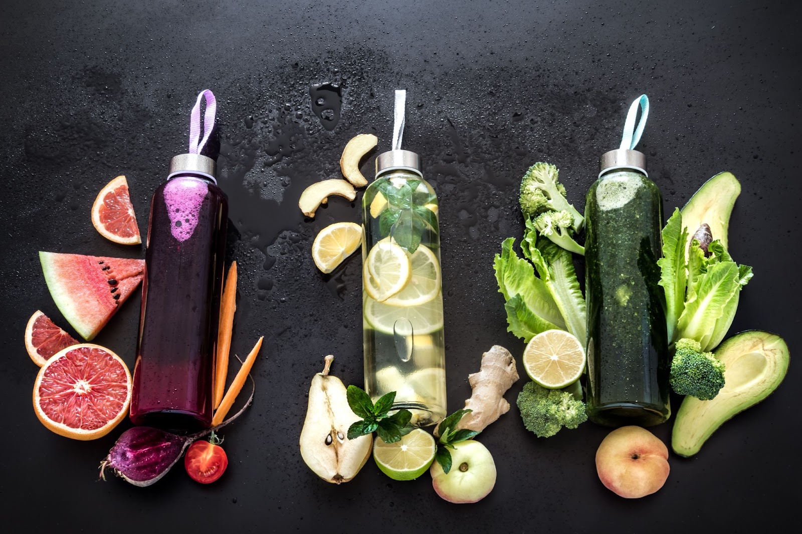 A flat lay of three different natural detox smoothie bottles on a black background, showcasing vibrant colors and fresh ingredients from a top-down view.