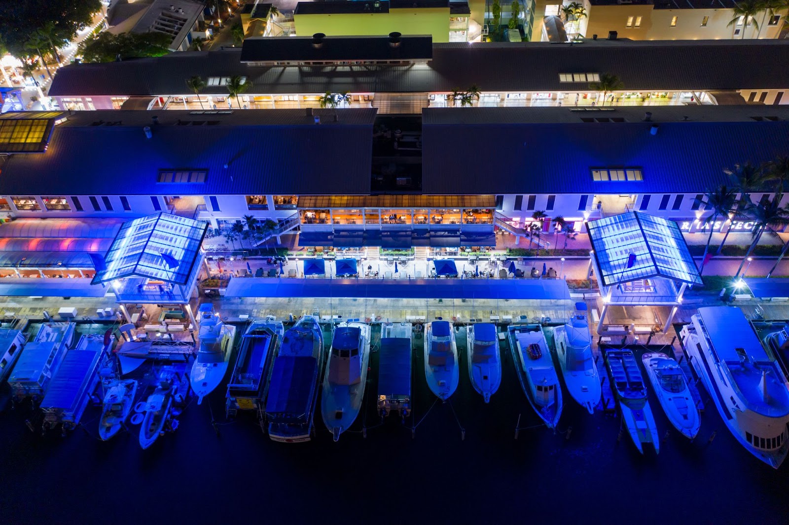 Aerial nighttime view of Bayside Marketplace in Miami, showing illuminated buildings, outdoor dining areas and a marina lined with yachts and boats under vivid blue lighting.