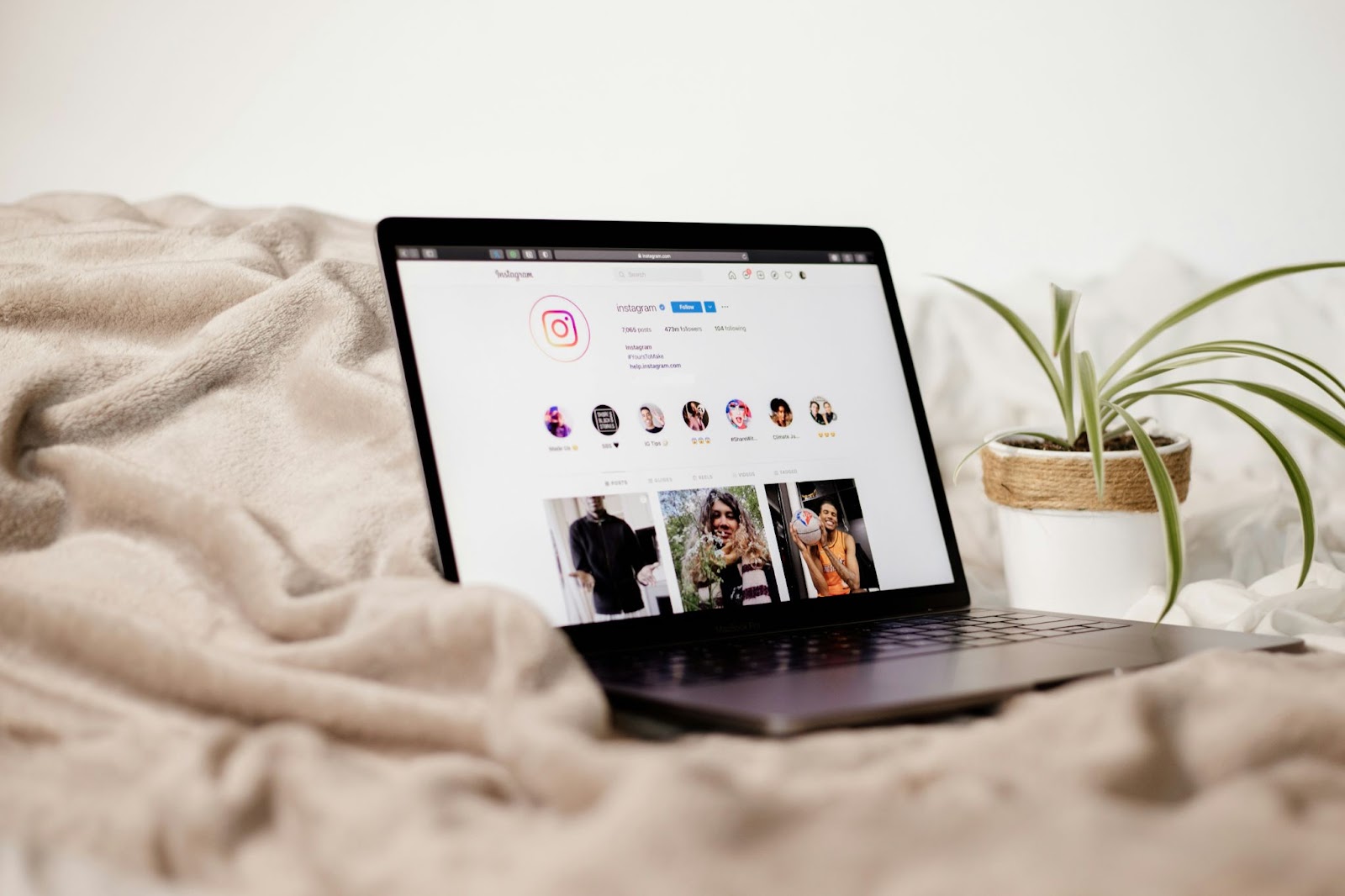 Laptop on a bed displaying an Instagram profile, next to a plant