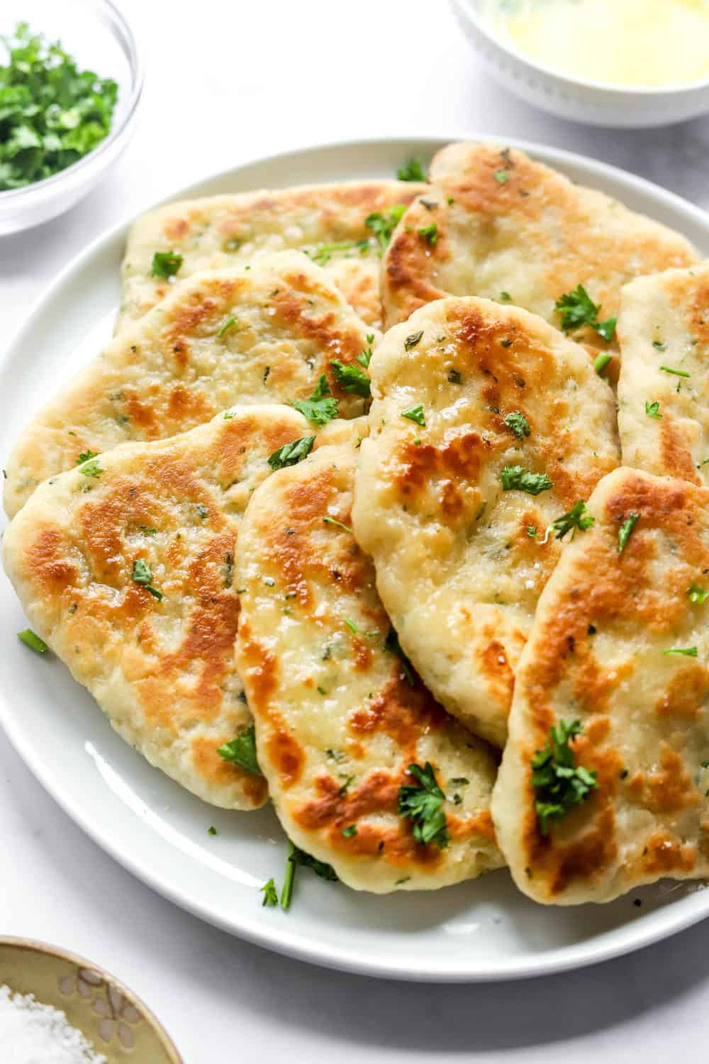 Layered naan flatbreads on a white plate topped with chopped green herbs and sea salt with more herbs behind it.