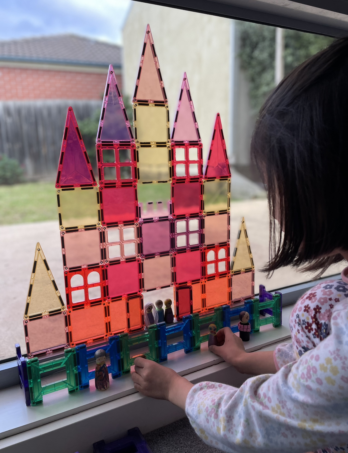 A toddler builds a warm-colored magnetic tile castle on the window, alongside a cool-colored fence. The child uses peg dolls for storytelling, engaging in imaginative play with the vibrant structures, highlighting the open-endedness of magnetic tiles that encourage creativity, problem-solving, and endless exploration.