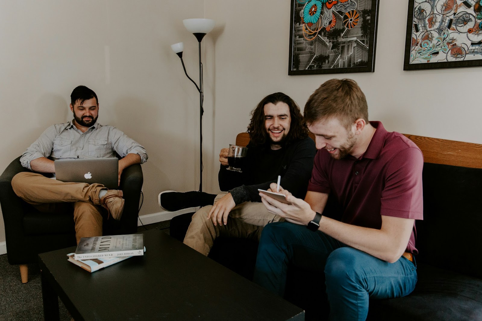 Three men lounging on a couch, laptops open and coffee cups in hand, deep in conversation or work.