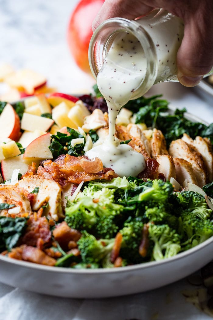 A photo of a kale salad loaded with apples, bacon, cranberries and smoked chicken topped with poppy seed dressing being poured on top.