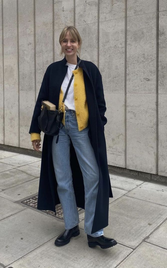 wanita memakai celana jeans biru, kaos putih, cardigan kuning, long coat navy, dan tas&nbsp;