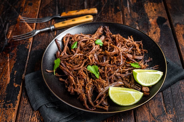 A plate of flavorful Carnitas garnished with fresh cilantro and lime wedges, served on a dark plate with rustic wooden utensils in the background