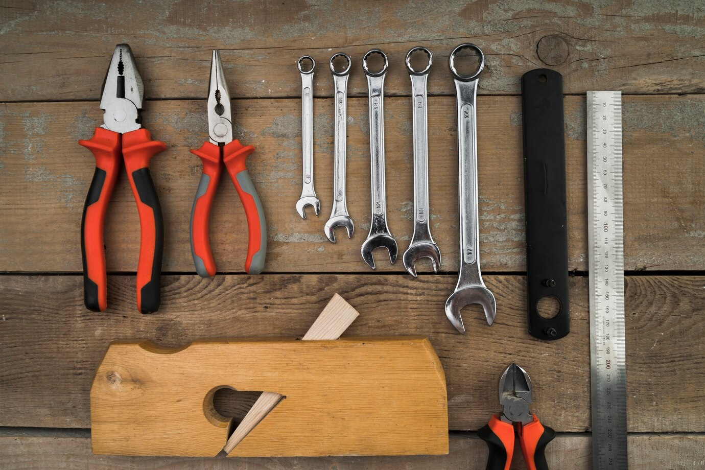 an assortment of tools neatly arranged on a wooden surface. The tools include pliers with red and black handles, a set of open-end and box-end wrenches of various sizes, a carpenter's wooden hand plane, a ruler, and other utility tools