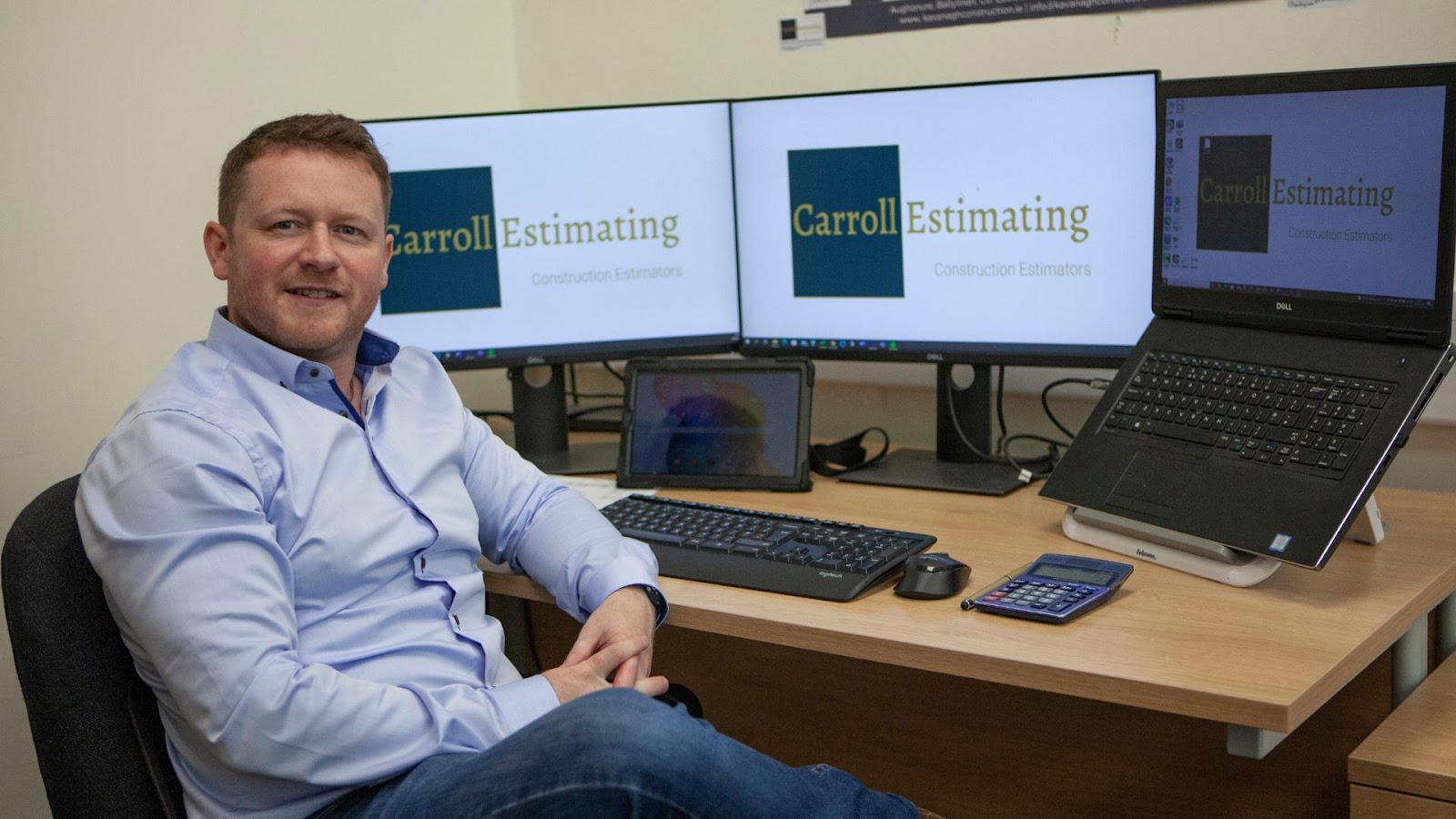 Paddy sitting infront of computer screens with ocmpany logo. 