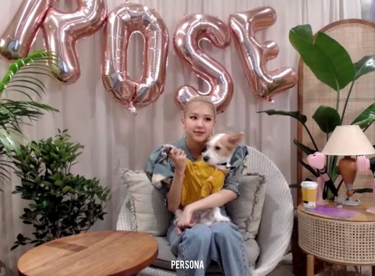 This contains an image of BLACKPINK Rosé sitting in a chair holding a small dog with the word rose on it