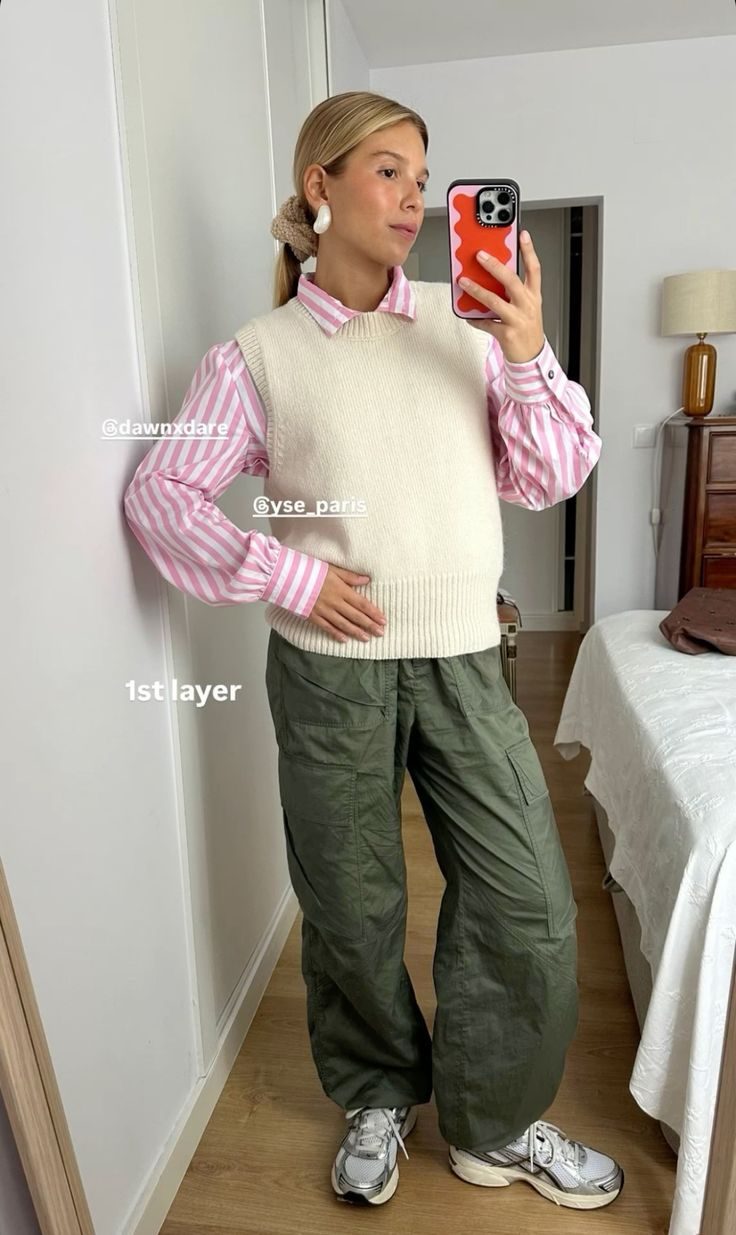 wanita memakai celana hijau, kemeja bergaris pink, sweater vest krem berfoto di depan cermin