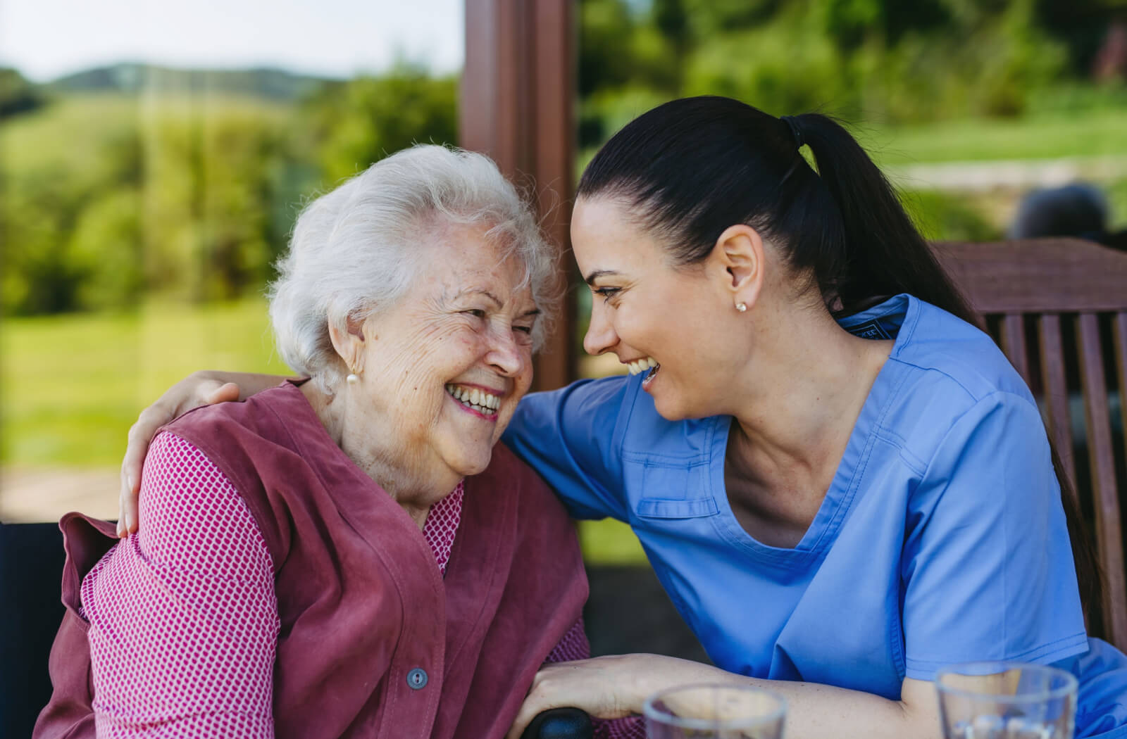 A caregiver and an older adult with dementia sitting near a window of a memory care community laughing with one another.