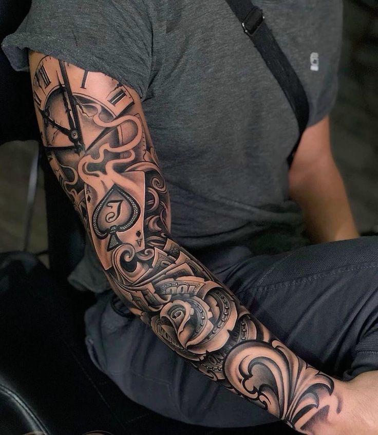 Detailed sleeve tattoo with clock, ace of spades, and ornate designs on a person's arm, wearing a gray shirt and black strap.