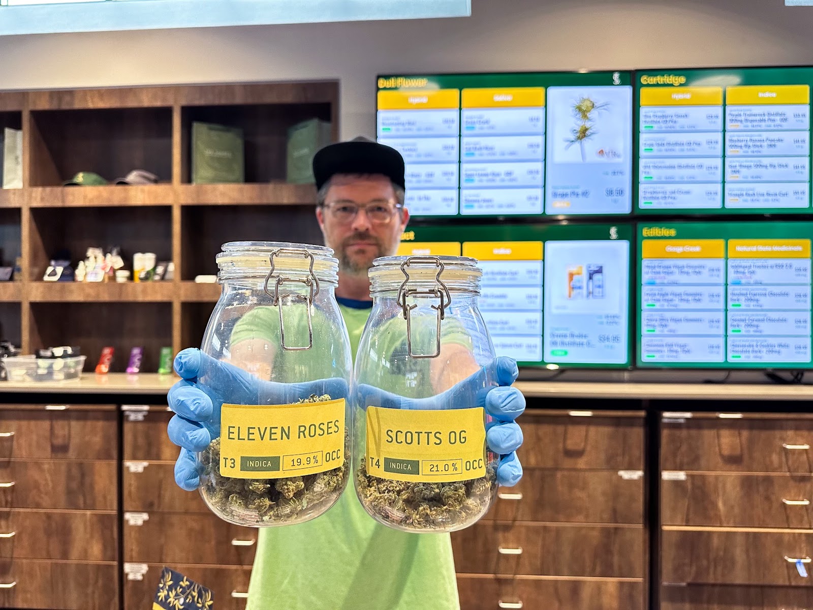 Budtender at The Source cannabis dispensary located in Rogers, Arkansas, holds up jars of popular cannabis indica flower strains, Scott’s OG and Eleven Roses by Osage Creek Cultivation.