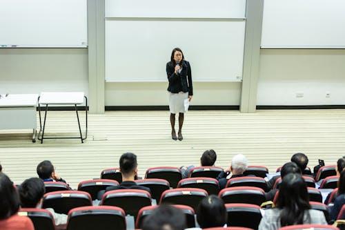 Free Female speaker presenting to an audience in a modern auditorium setting. Stock Photo