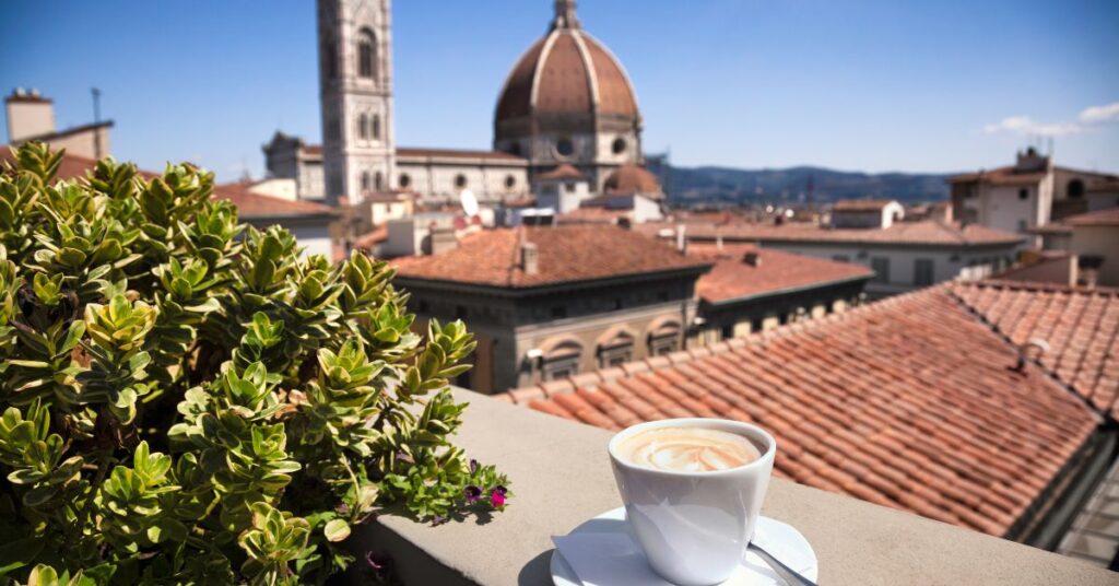 A latte in a white mug on a white plate on a patio overlooking a town in Italy with red tiled roofs.