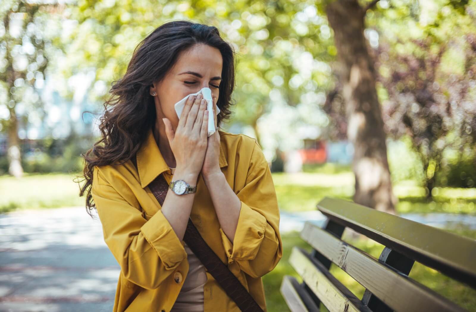 A patient sitting on a bench outside blows their nose as they deal with the effects of seasonal allergies.