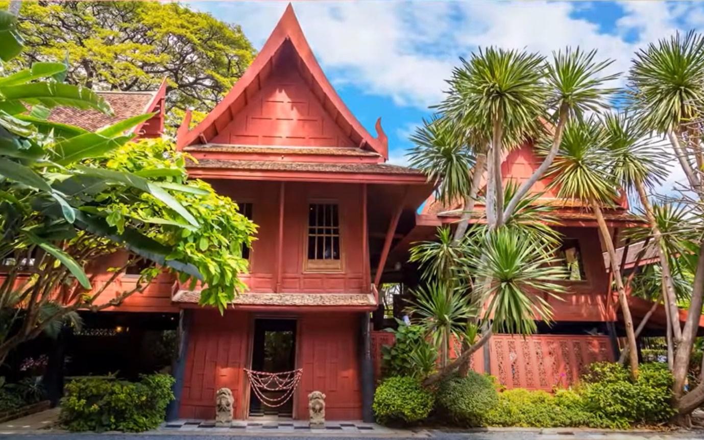 A red house with trees and bushes with Jim Thompson House in the background

Description automatically generated