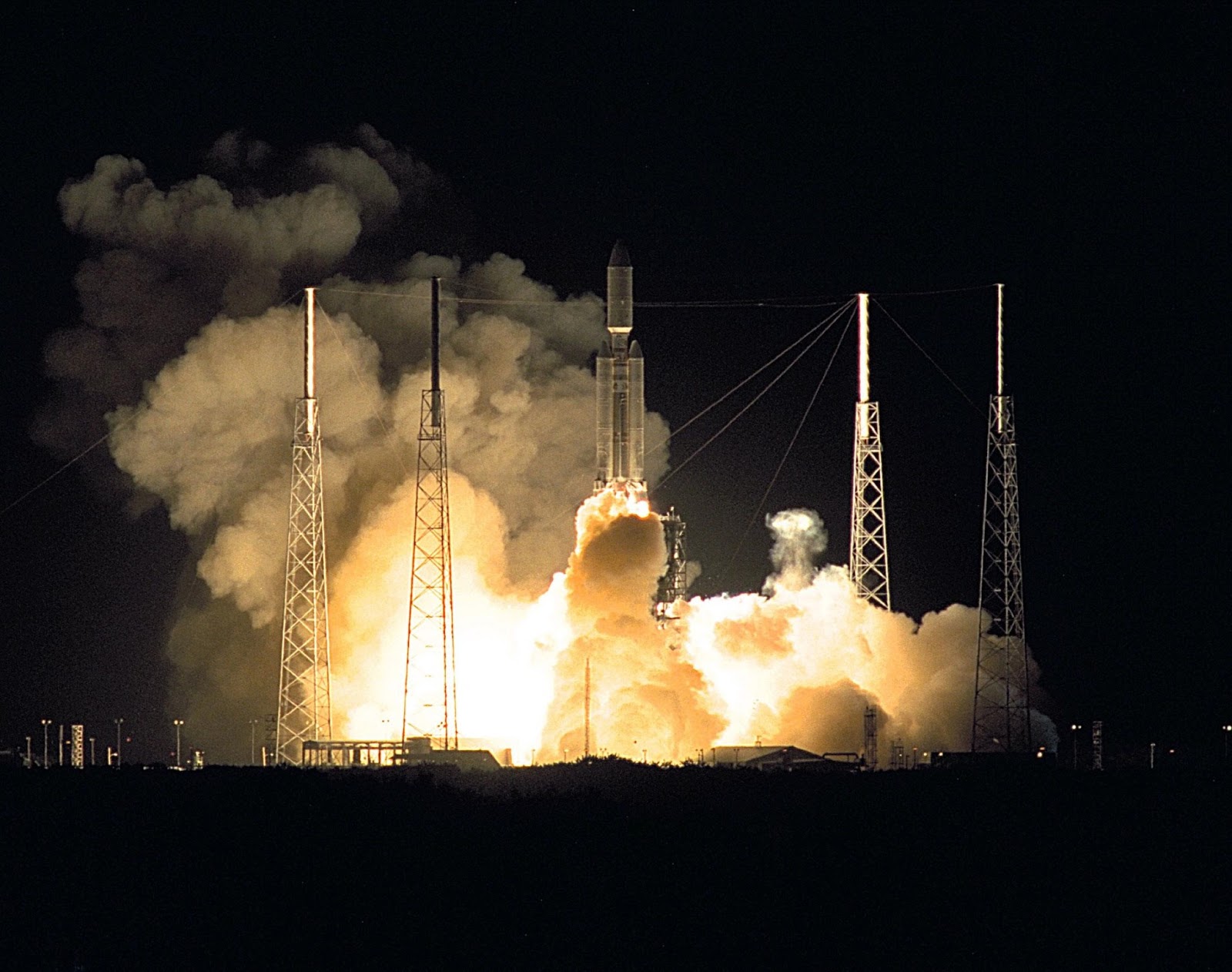 Surrounded by darkness, a large rocket blasts off from a launchpad as orange-white smoke billows out from underneath it.