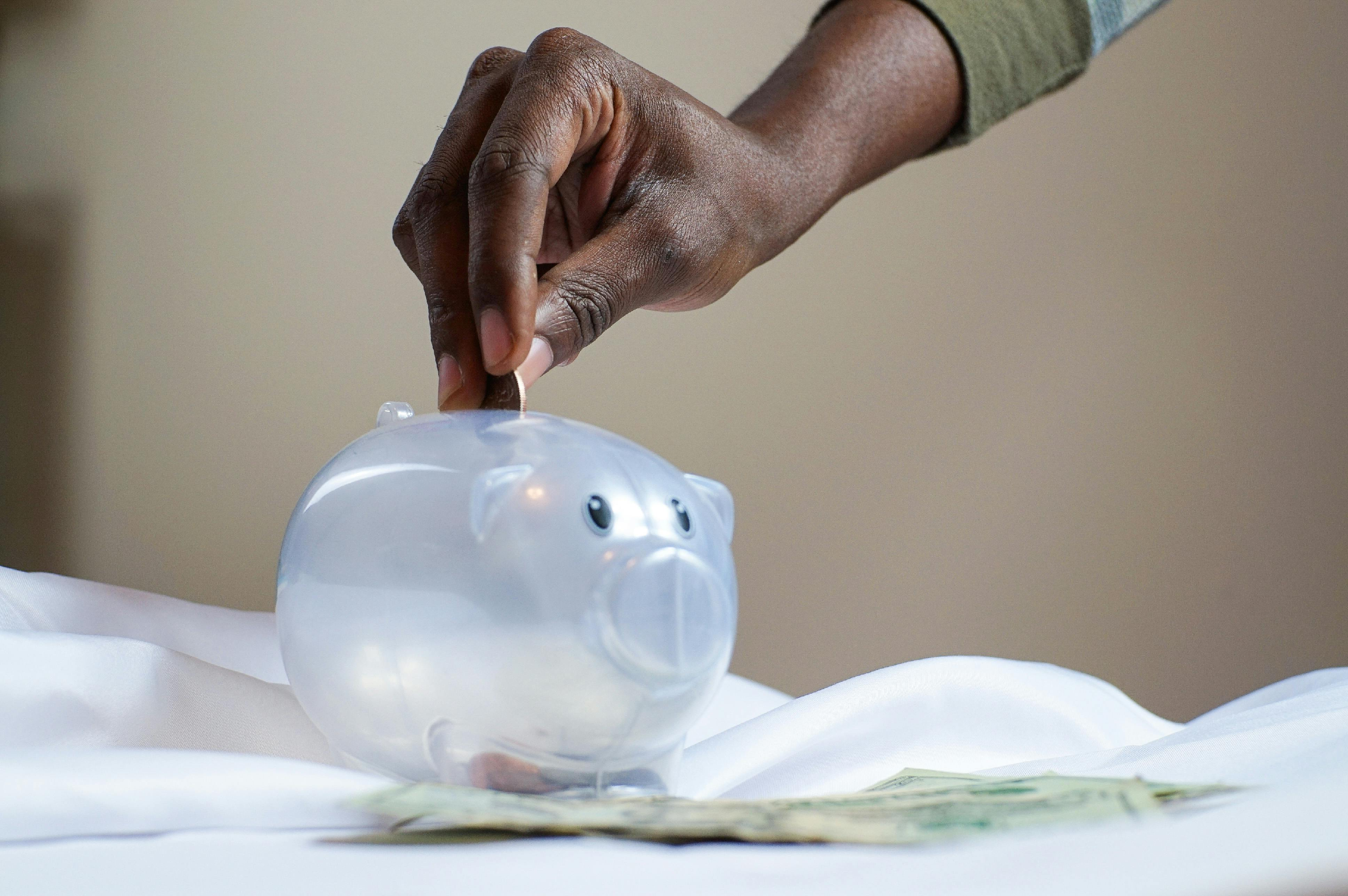 A hand putting a coin into a clear piggy bankAI-generated content may be incorrect.
