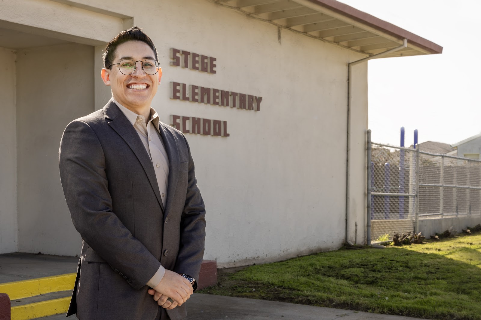 A man with glasses and a gray suit stands in front of an elementary school.