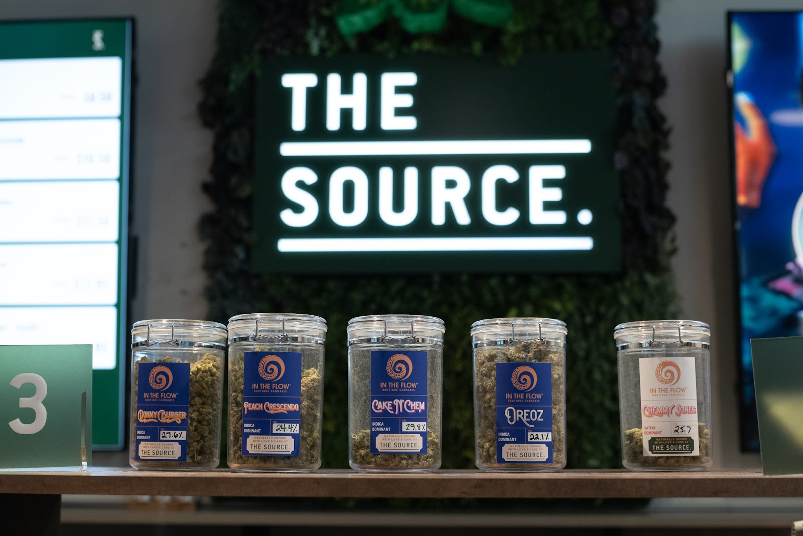 Five jars of cannabis sit on a shelf in front of a bright sign that says "The Source." The cannabis strains in the jar are brightly labeled: Donny Burger, Peach Crescendo, Cake N' Chem, Oreoz, and Chemmy Jones, all produced by In The Flow boutique cannabis. 