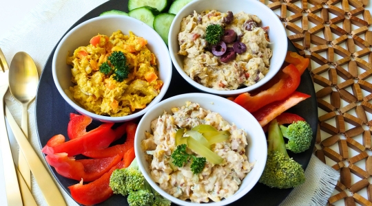 A platter featuring three bowls of different tuna spreads, garnished with vegetables and served with sliced cucumbers, red peppers, and broccoli.