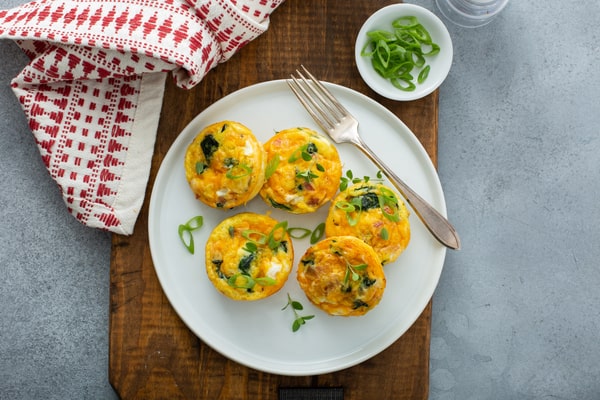 A plate of savory egg bites topped with chopped green onions, served with a fork and napkin on a wooden board