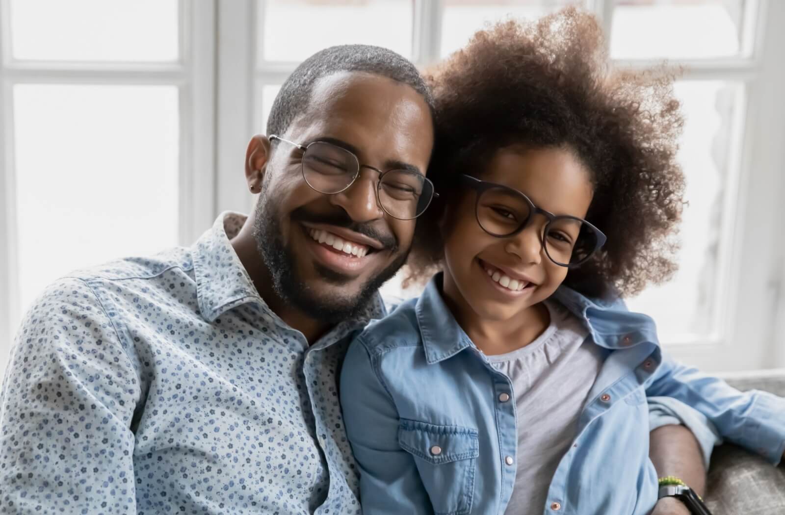  A parent and child smile together, wearing their new eyeglasses and seated against a window