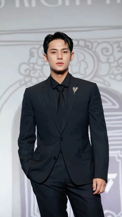 This contain SEVENTEEN's Mingyu in a black suit and tie standing on a runway with his hands in his pockets