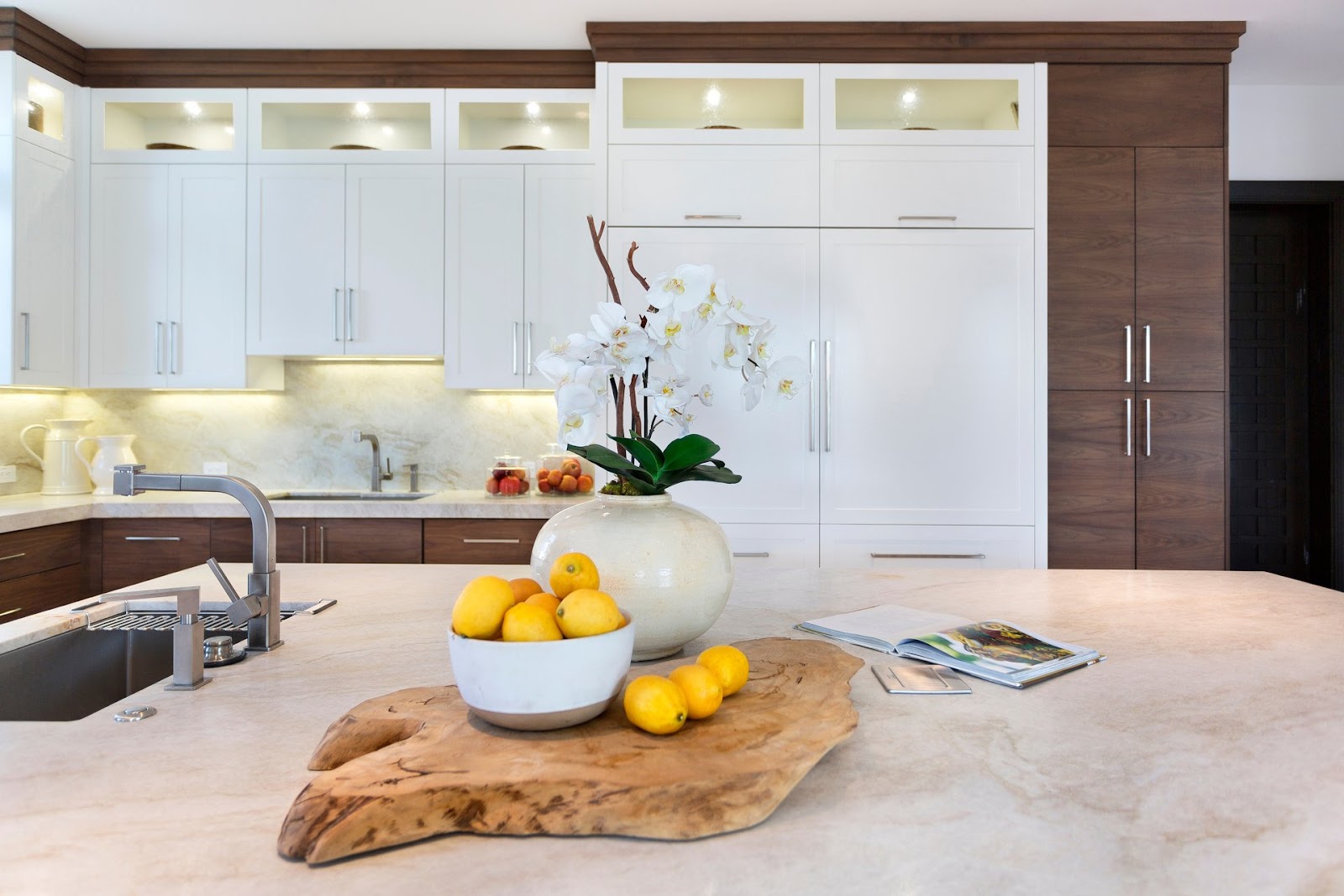 A modern kitchen featuring white cabinets, a marble countertop, a vase with white orchids, and a bowl of lemons on a wooden platter.