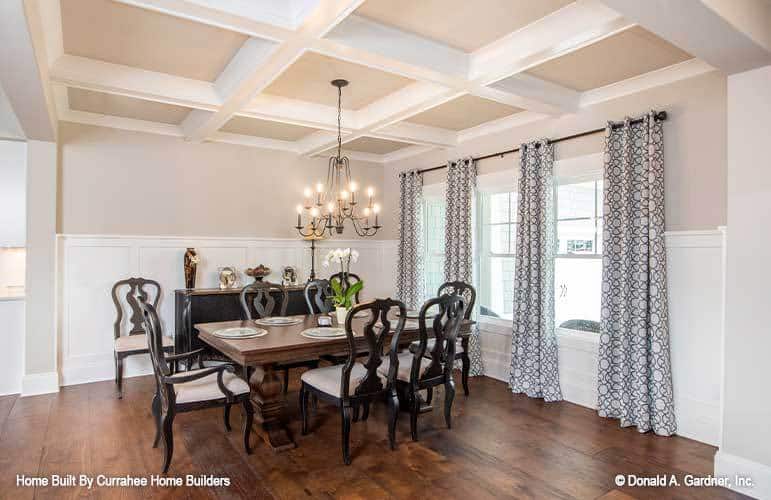 The formal dining room has a coffered ceiling, wainscoted walls, a 3-seater dining set, and a trio of windows dressed in patterned drapes.