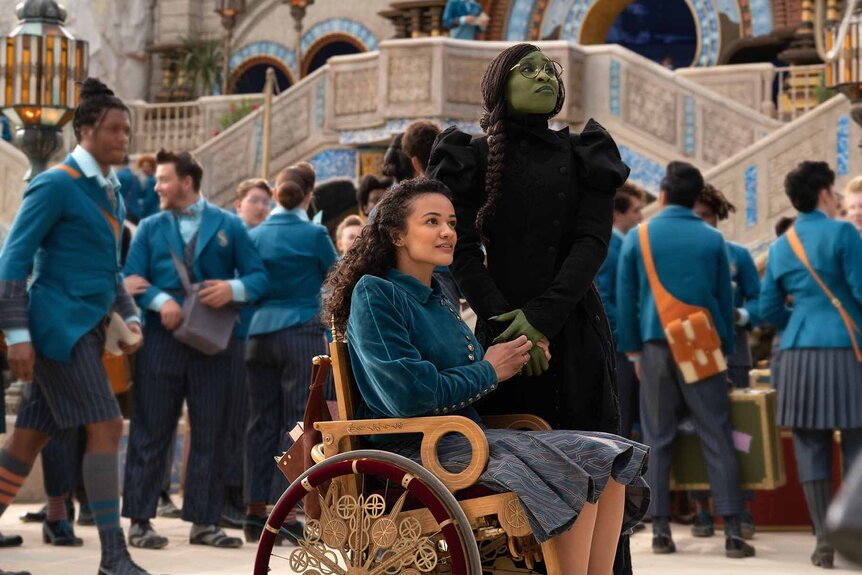 Elphaba con piel verde y vestimenta negra junto Nessarose en silla de ruedas, en un ambiente lleno de estudiantes con uniformes azules.