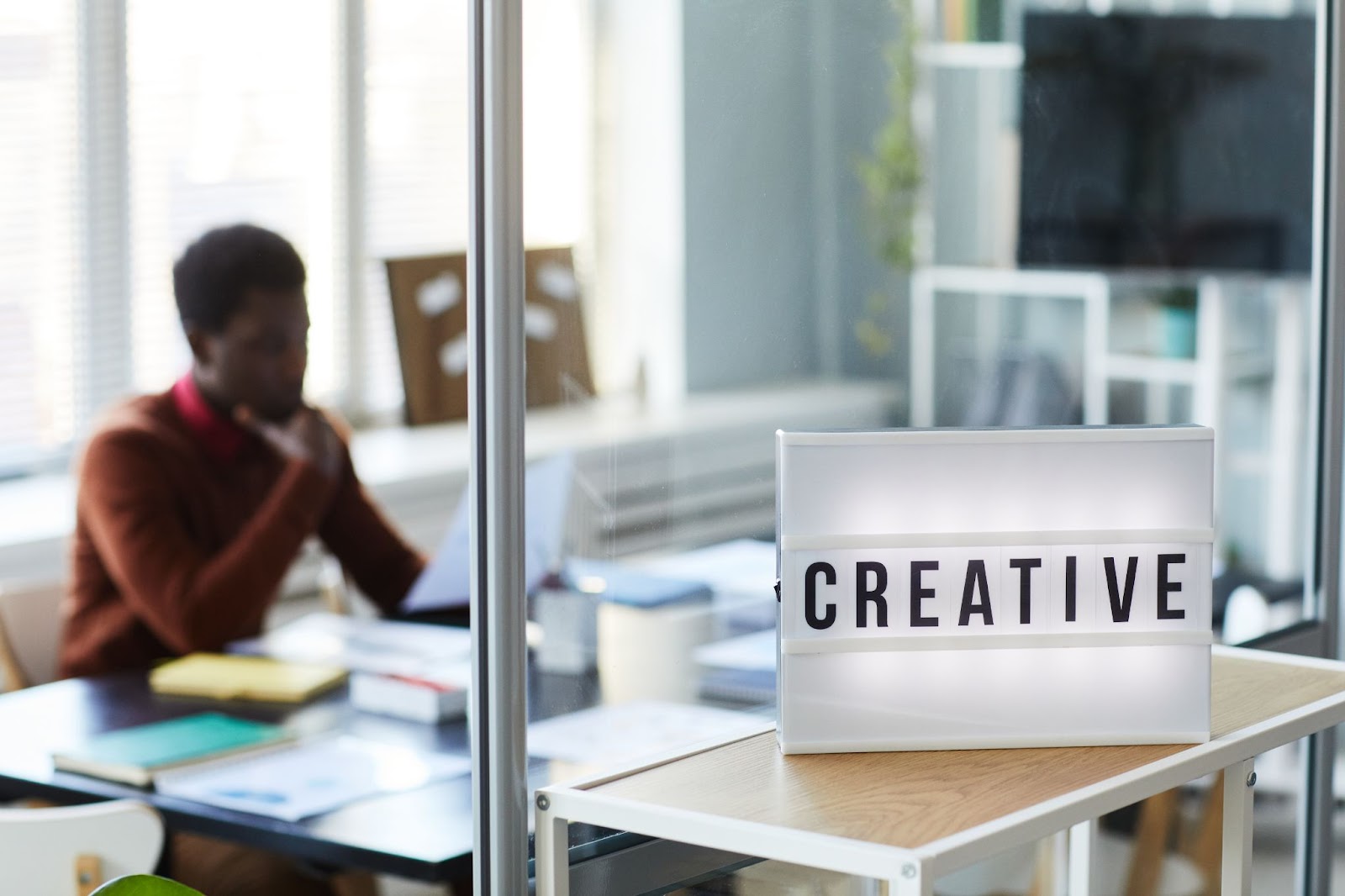 A sign with the word “creative” on it outside a glass office with a man inside. 